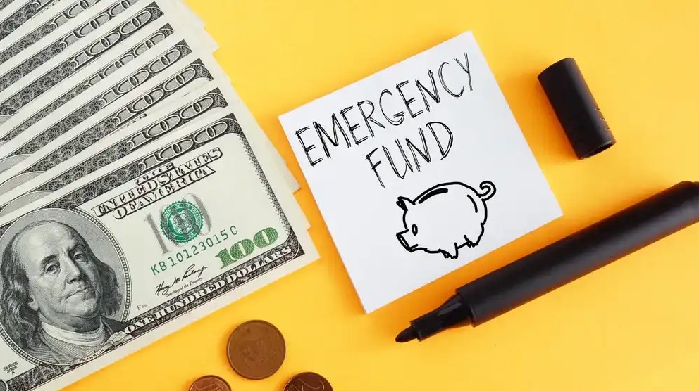 $100 bills pile next to post it that says emergency fund